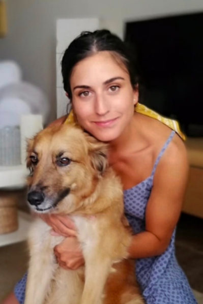 Woman in a blue patterned sleeveless dress sitting indoors and hugging a medium-sized tan and white dog, both looking toward the camera.